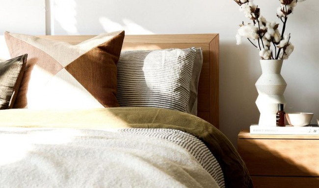 swath of sunlight shines across a well made bed and bedside table with a vase of cotton plants